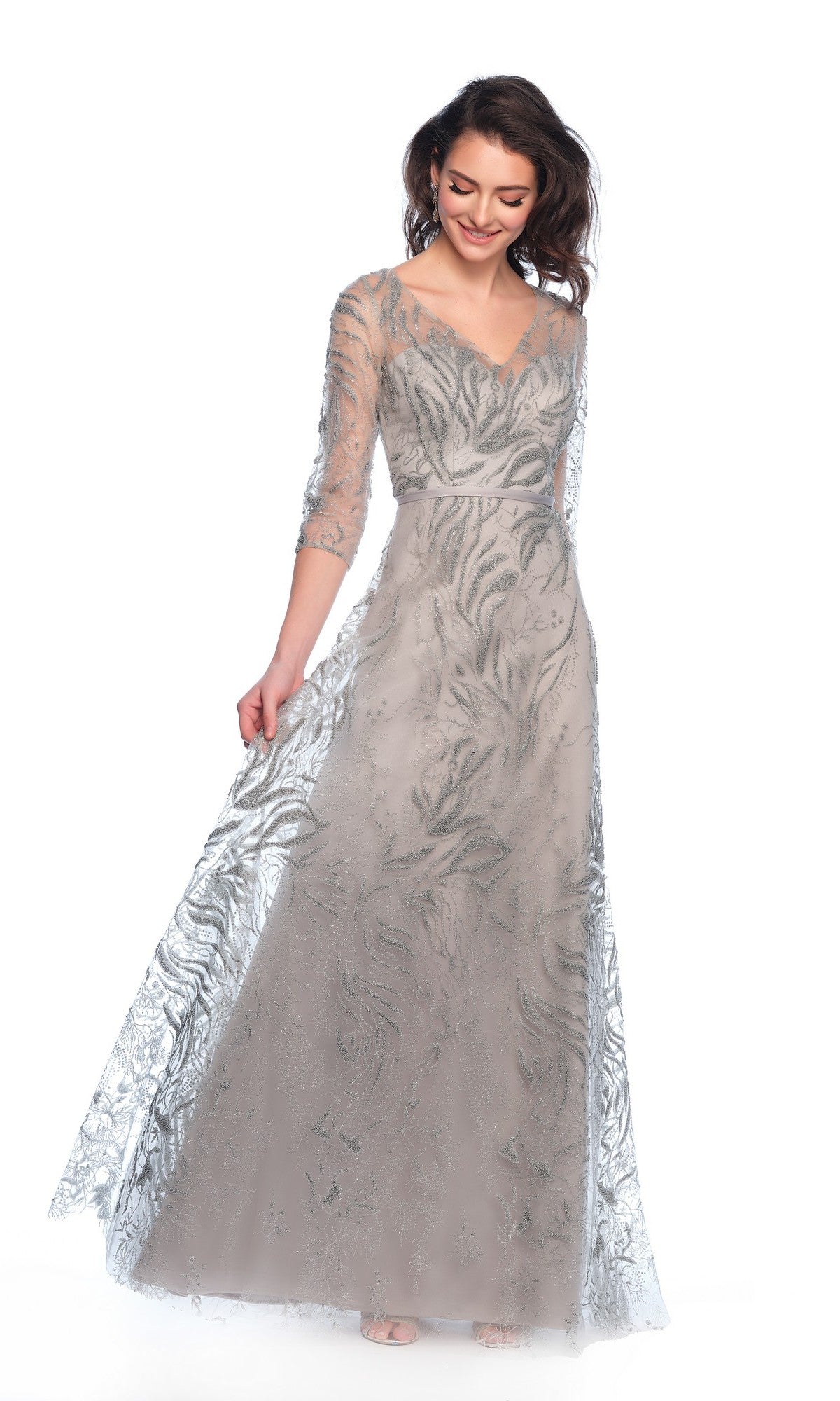 A woman stands against a white background wearing the Long Formal Dress 11606 by Dave and Johnny—an elegant silver A-line gown with sheer sleeves, intricate leaf patterns, and a subtle sweetheart neckline.